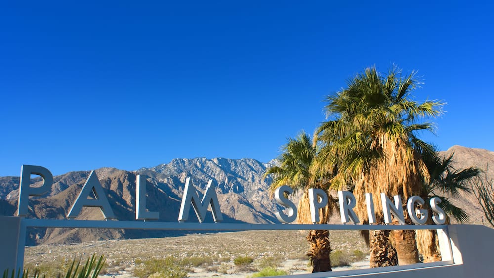 A sign reading "PALM SPRINGS" with palm trees and mountains in the background.