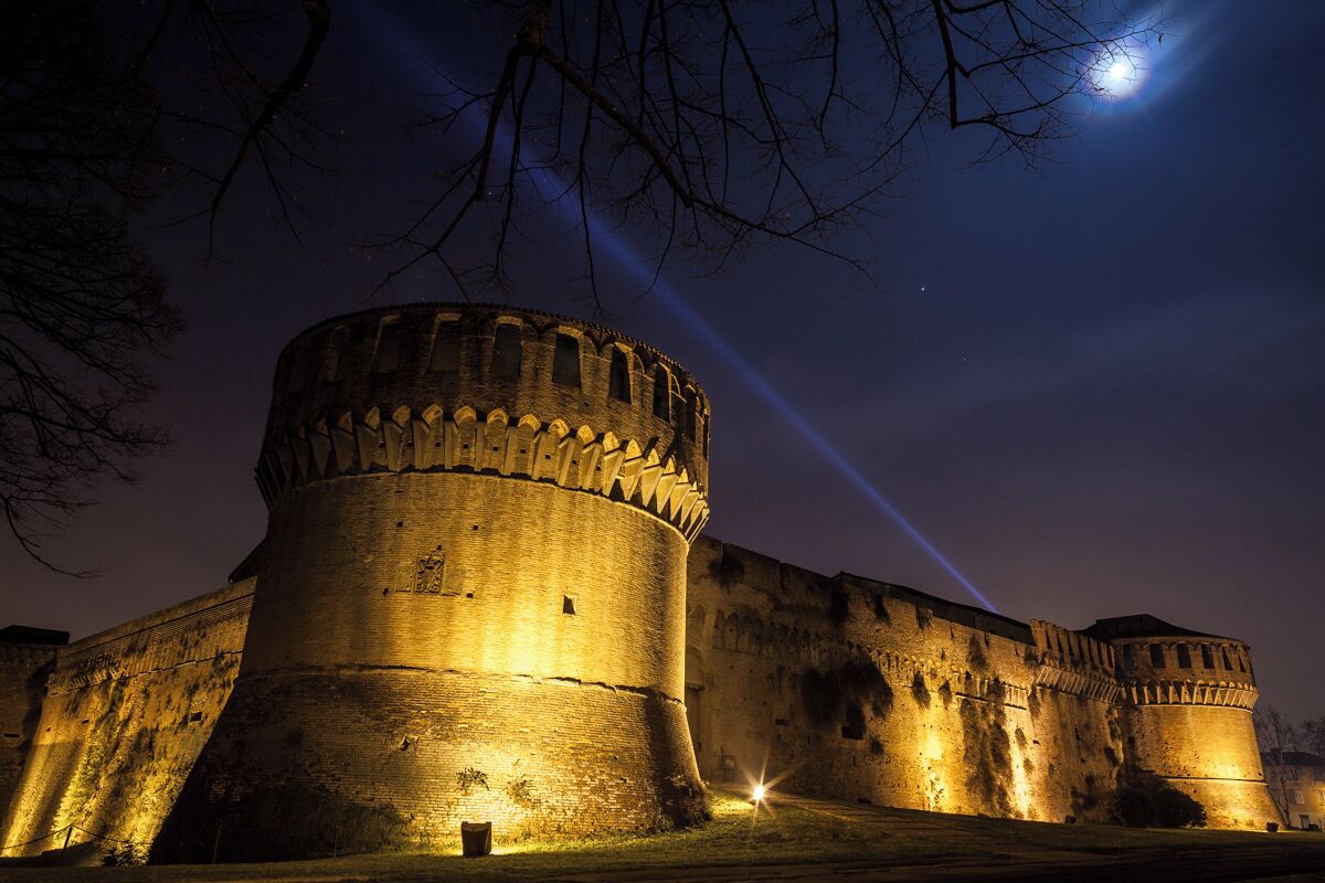 A historic stone castle with a large round tower, illuminated by artificial lighting, under a night sky with a visible moon.