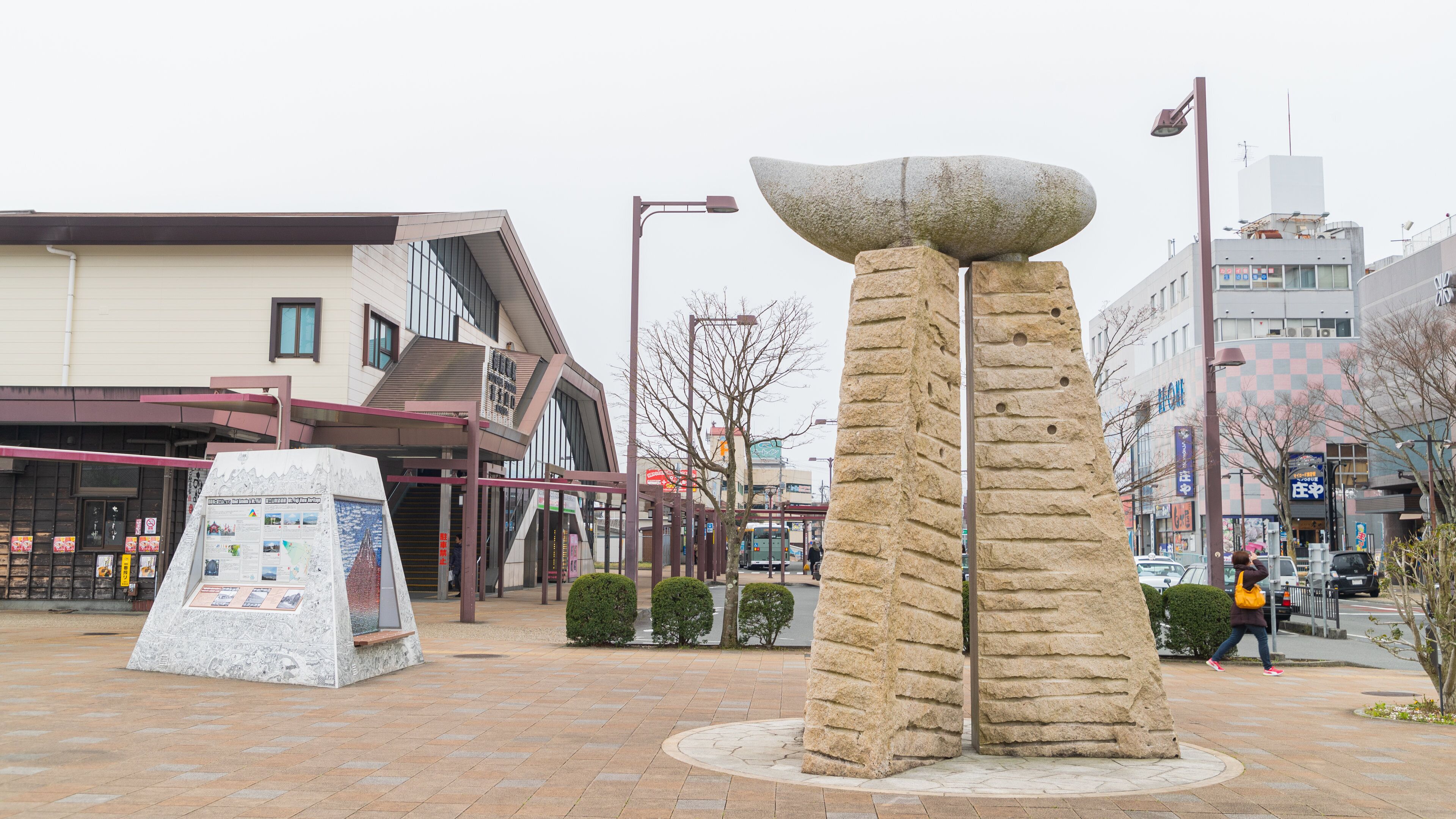 Una escultura de piedra en una plaza urbana, con una parada de autobús y edificios comerciales al fondo.