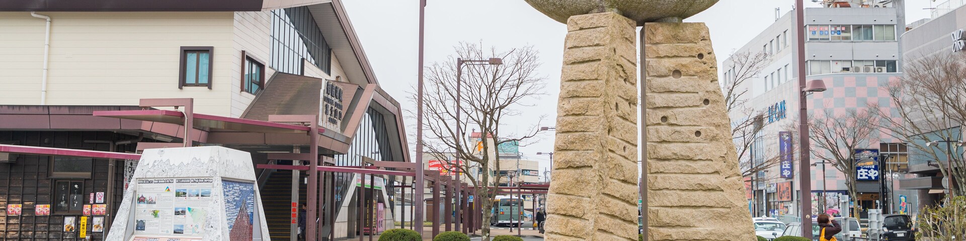 Una escultura de piedra en una plaza urbana, con una parada de autobús y edificios comerciales al fondo.