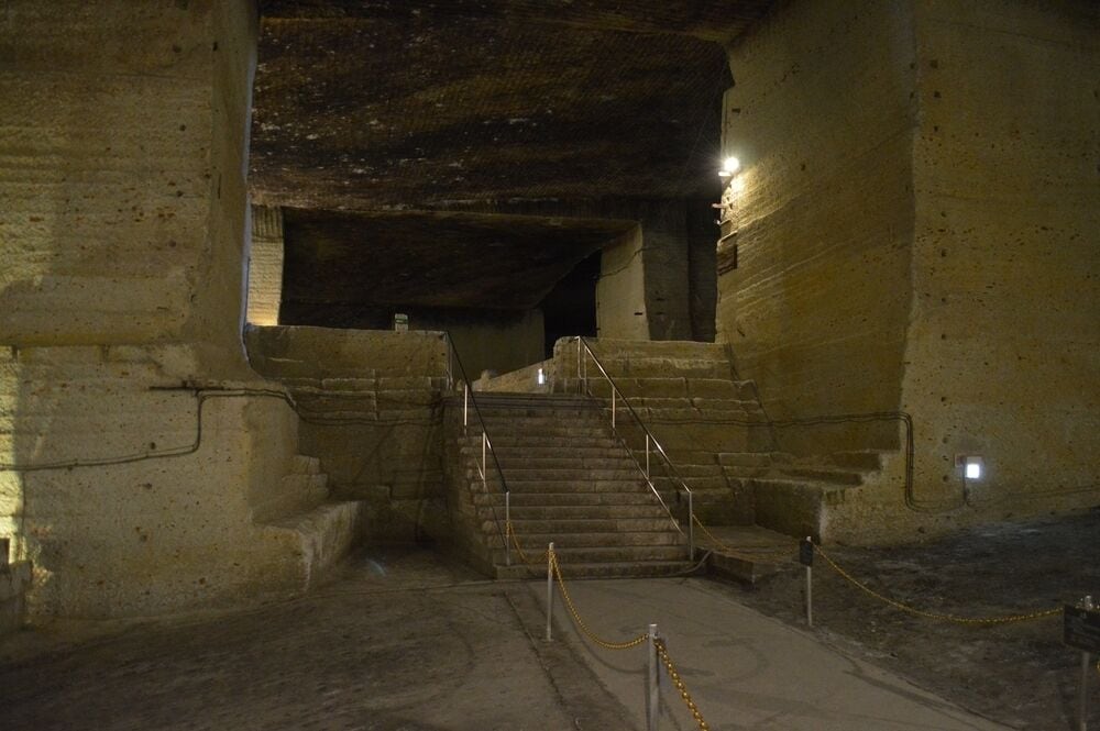 Una escalera de piedra tenuemente iluminada que desciende a un nivel inferior, rodeada de muros de piedra y un suelo de hormigón.