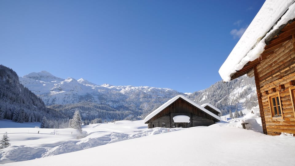 A wooden cabin in a snowy mountain valley with snow-covered trees and a clear blue sky.
