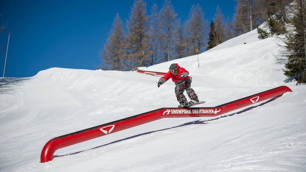 Un snowboardeur effectuant une figure sur une barre rouge dans un paysage montagneux enneigé.
