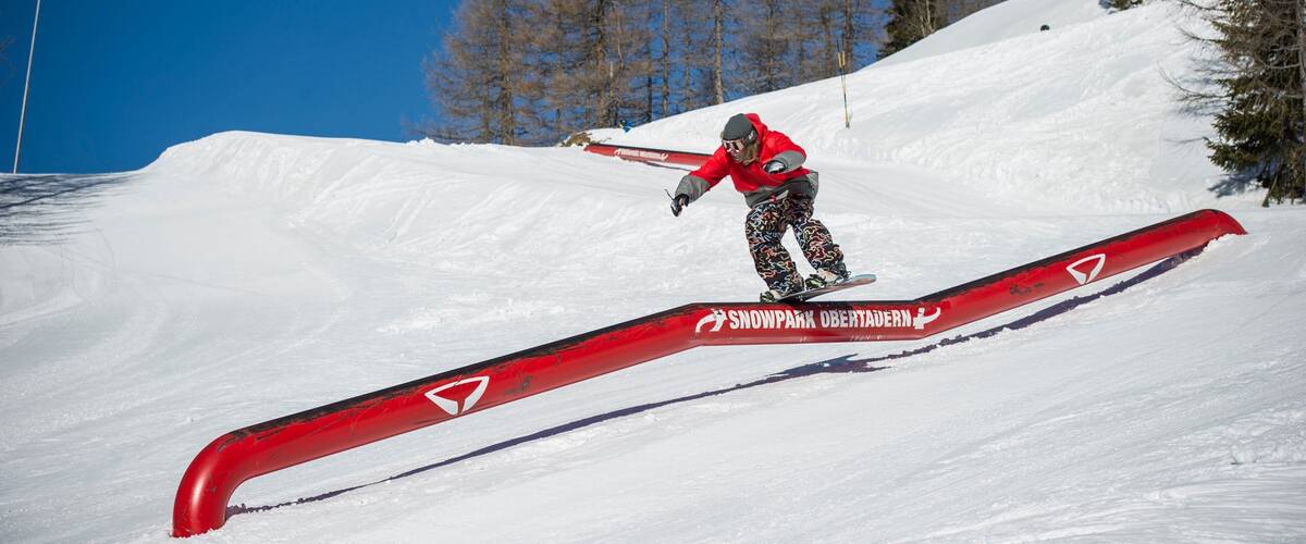 Un snowboardeur effectuant une figure sur une barre rouge dans un paysage montagneux enneigé.