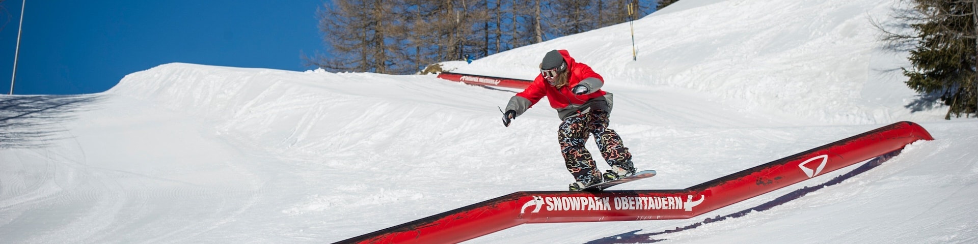 Un snowboarder realizando un truco sobre un riel rojo en un paisaje montañoso nevado.