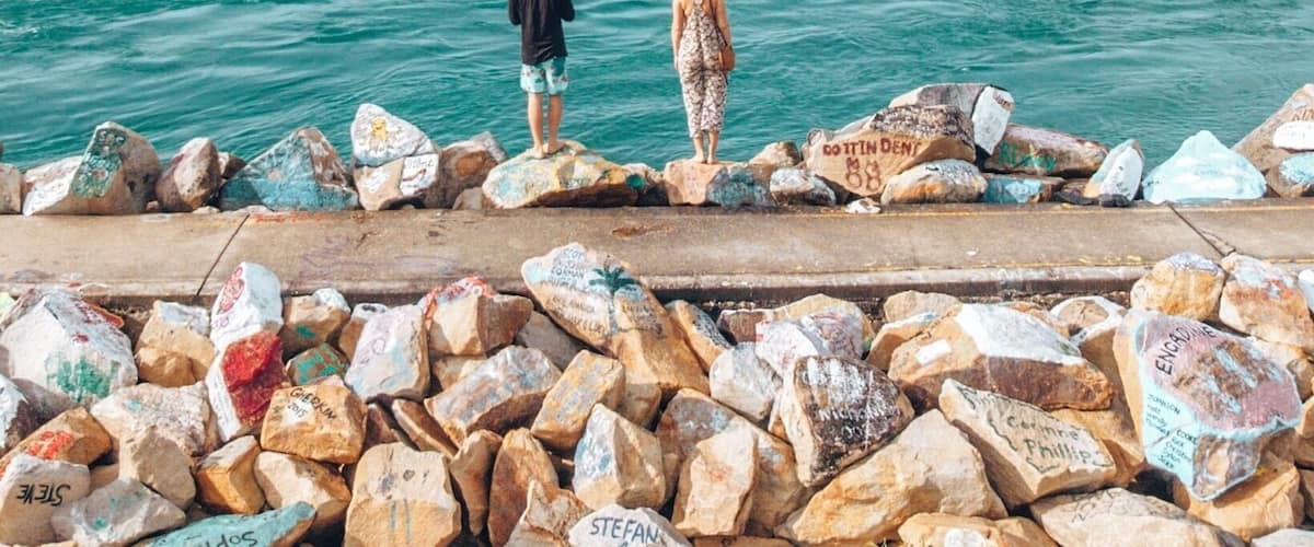 Two people standing on a rocky breakwater by a beach with a clear sky.