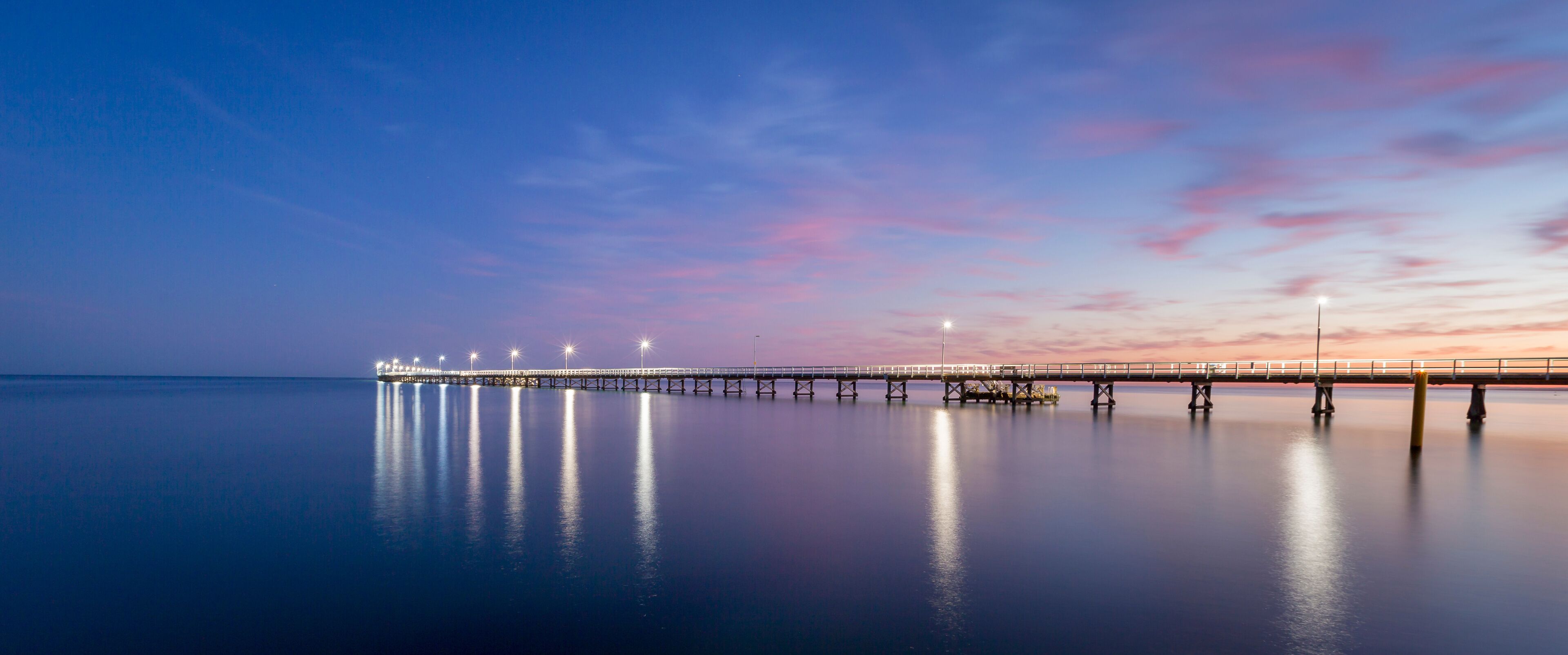 A long pier extending into calm waters, illuminated by lights, with a sunset sky.