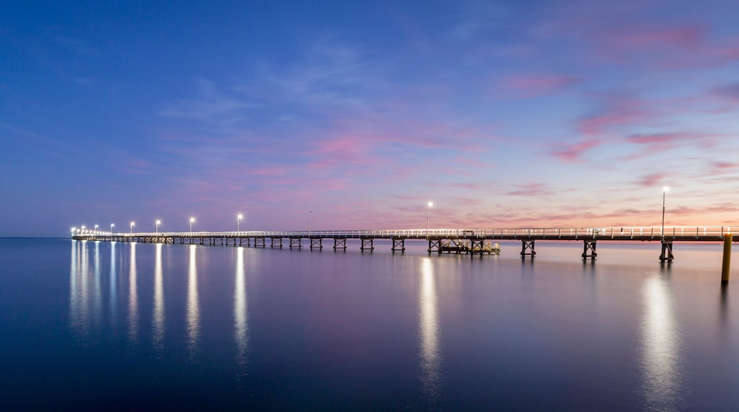 A long pier extending into calm waters, illuminated by lights, with a sunset sky.