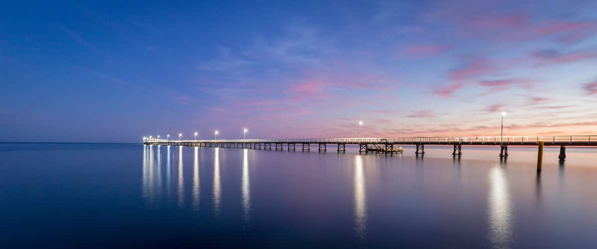 A long pier extending into calm waters, illuminated by lights, with a sunset sky.