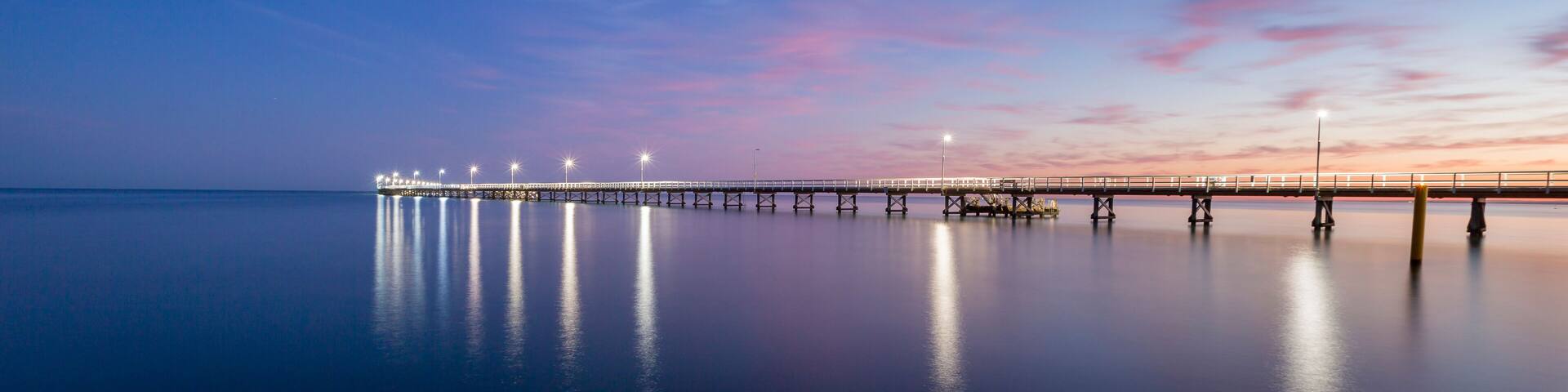 A long pier extending into calm waters, illuminated by lights, with a sunset sky.