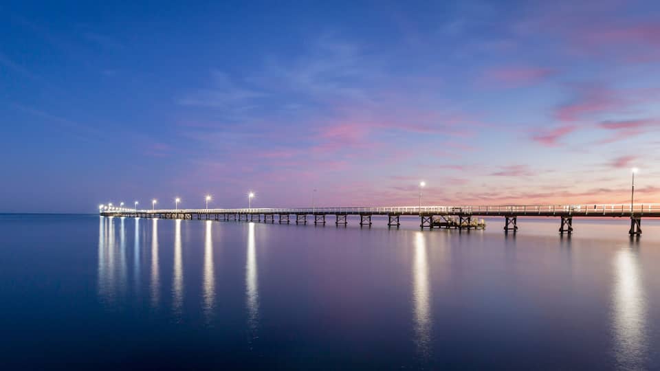 A long pier extending into calm waters, illuminated by lights, with a sunset sky.