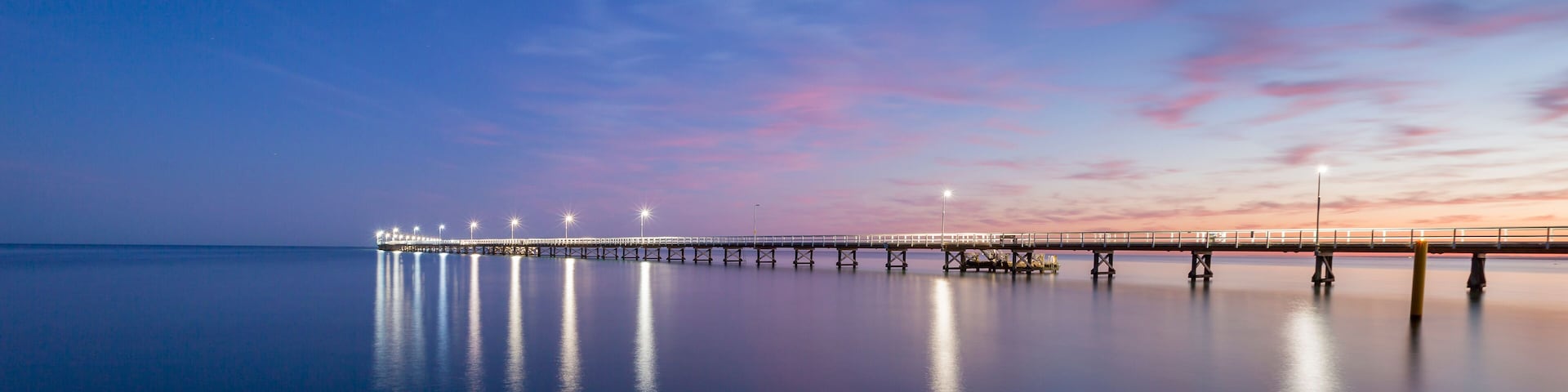A long pier extending into calm waters, illuminated by lights, with a sunset sky.