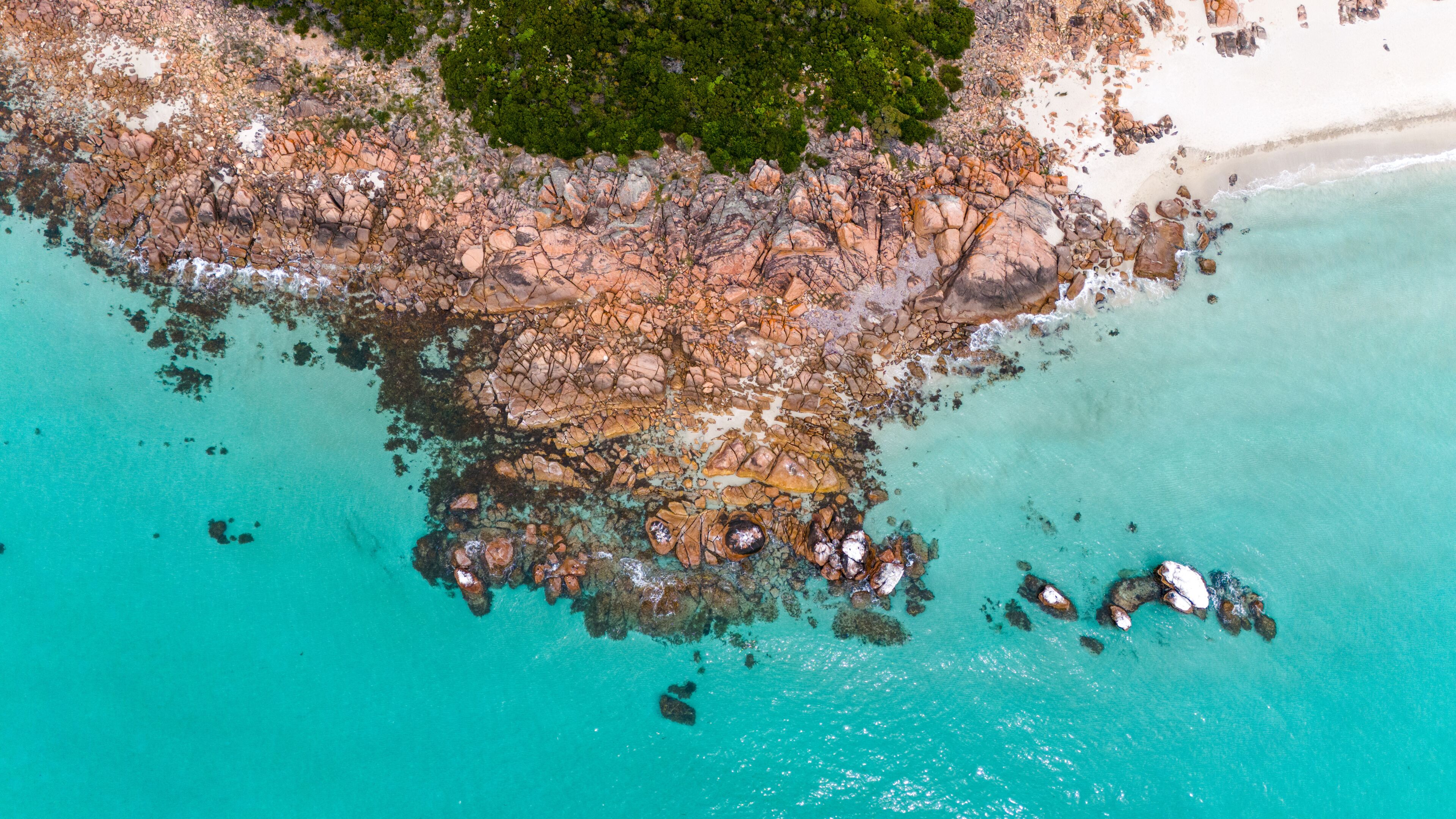 An aerial view of a rocky coastline with clear turquoise waters, a sandy beach, and dense green vegetation.