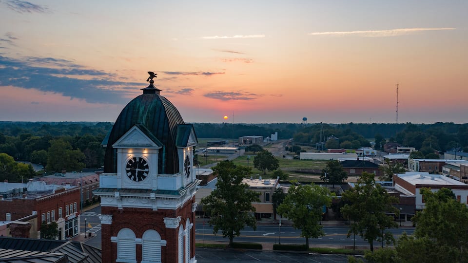 A clock tower with a dome, surrounded by a town with buildings and trees.