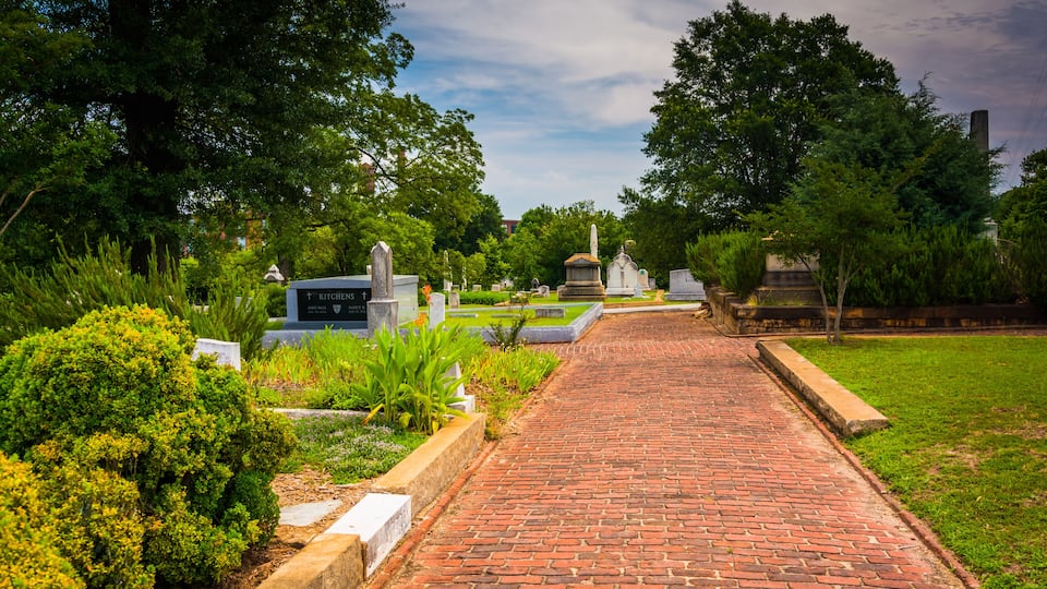 A brick pathway leading through a cemetery with various headstones and greenery.
