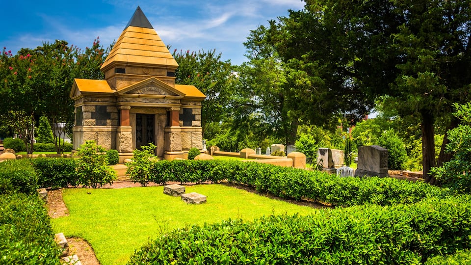 A small stone building with a pyramid-shaped roof surrounded by well-maintained hedges and trees.