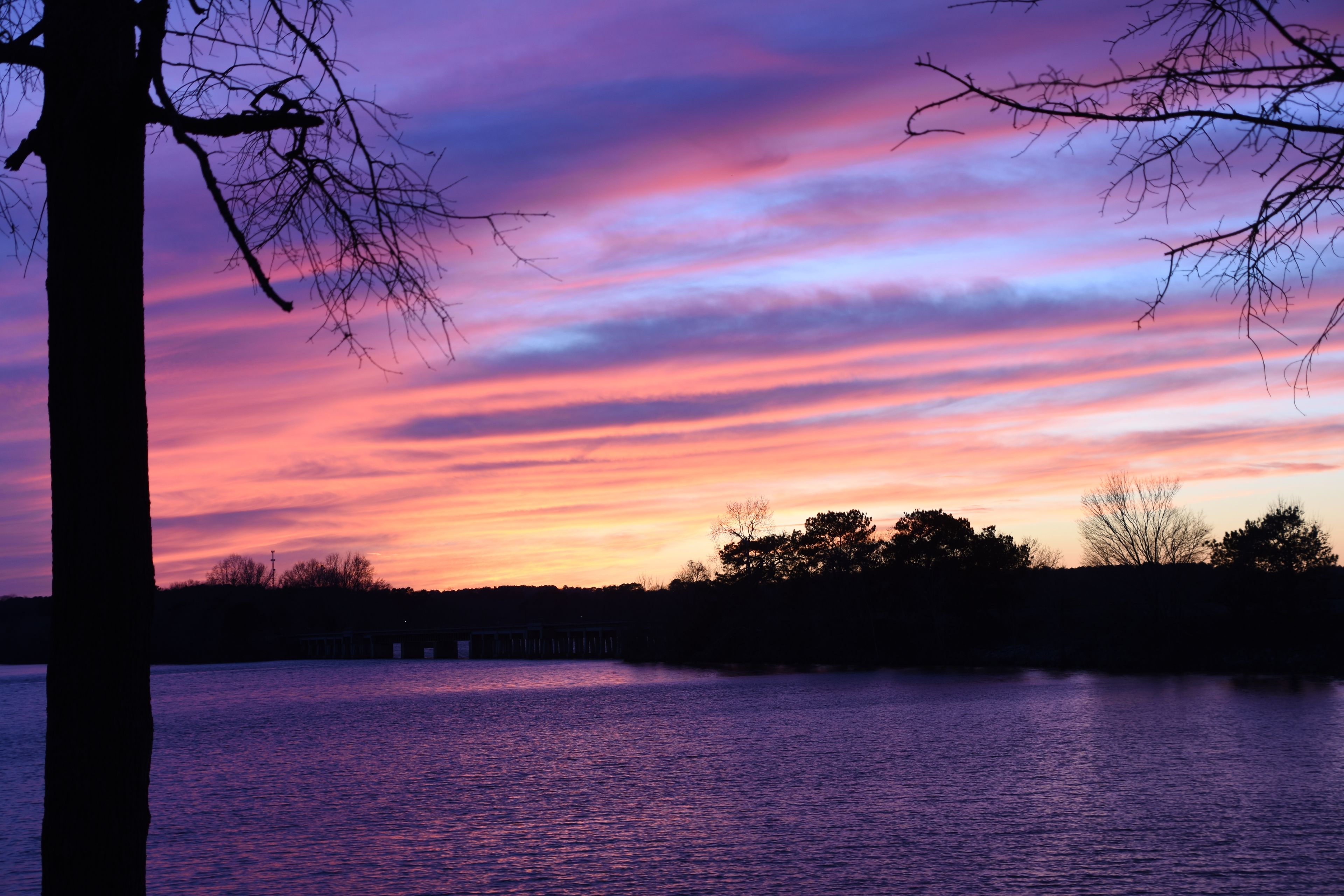 A sunset over a body of water with a bridge in the distance and silhouettes of trees.
