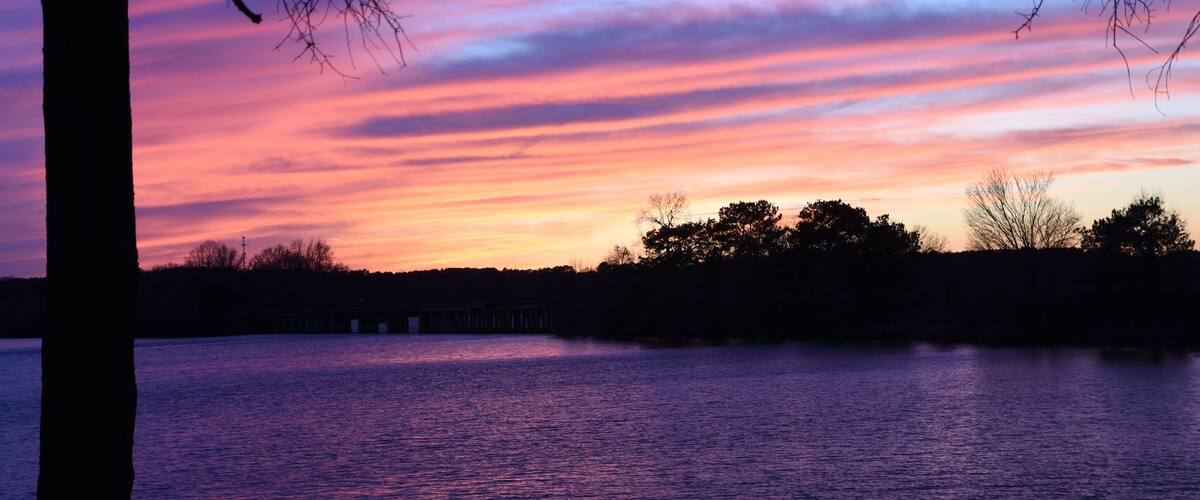 A sunset over a body of water with a bridge in the distance and silhouettes of trees.
