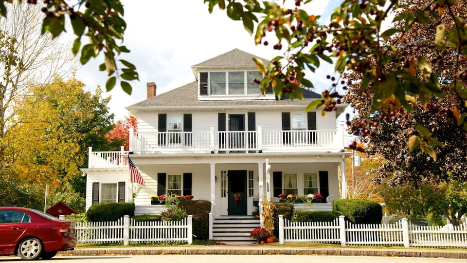 A two-story white house with a balcony, surrounded by a white picket fence, autumn foliage, and a red car parked in front.