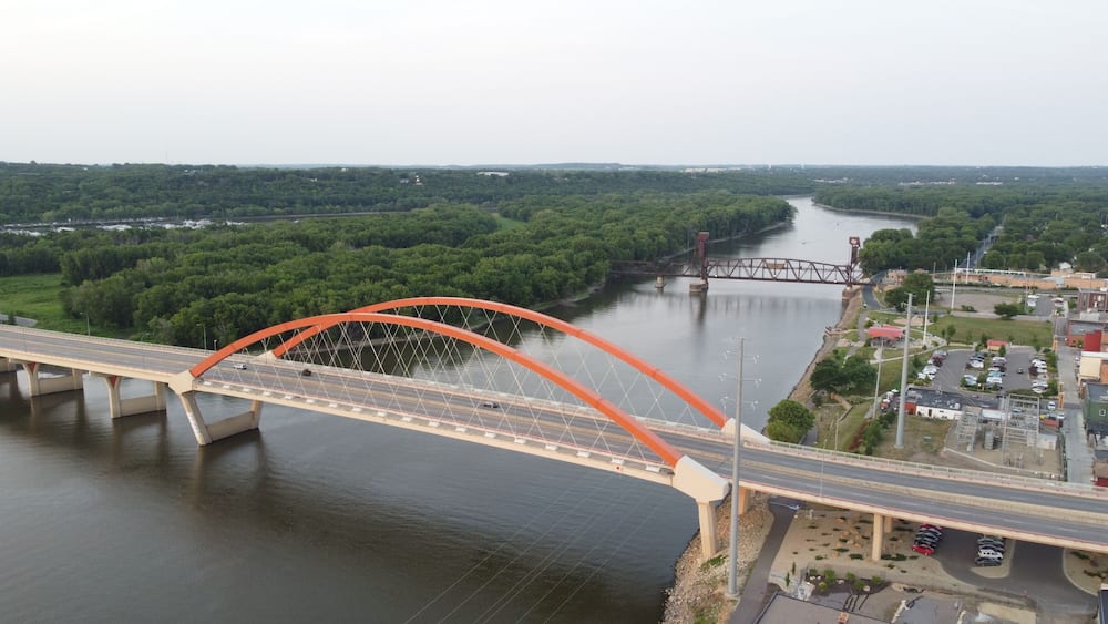 A large orange bridge spanning a river, surrounded by dense green forest and a small town with buildings and parked cars.