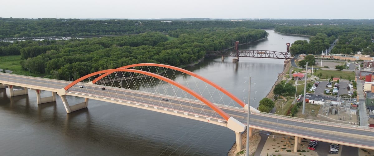 A large orange bridge spanning a river, surrounded by dense green forest and a small town with buildings and parked cars.