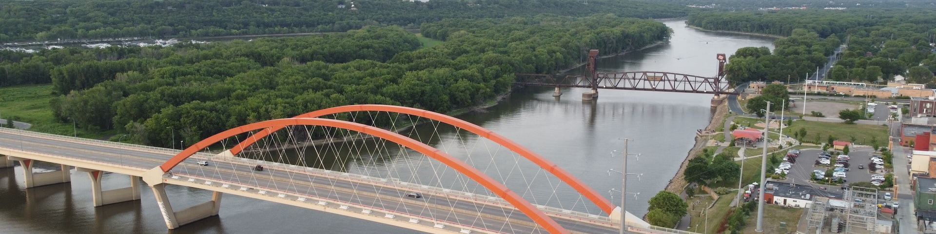 A large orange bridge spanning a river, surrounded by dense green forest and a small town with buildings and parked cars.