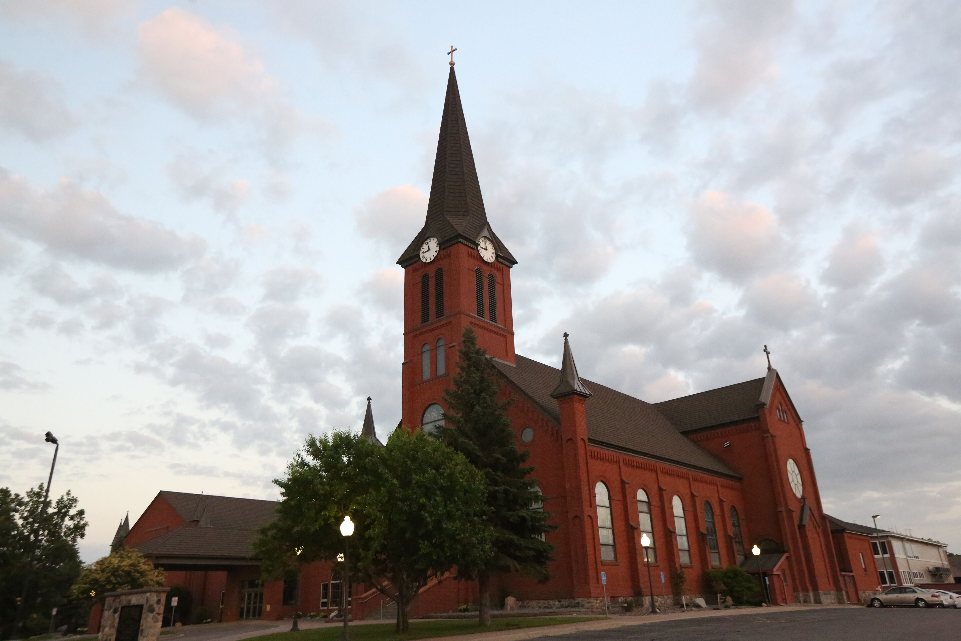 A red church with a tall steeple and two clocks, surrounded by trees and a cloudy sky.