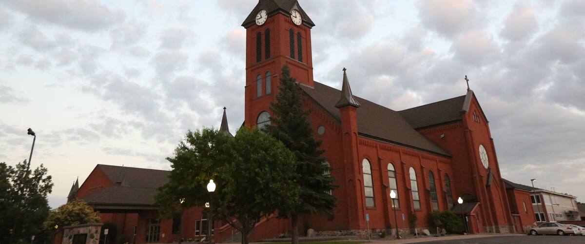 A red church with a tall steeple and two clocks, surrounded by trees and a cloudy sky.