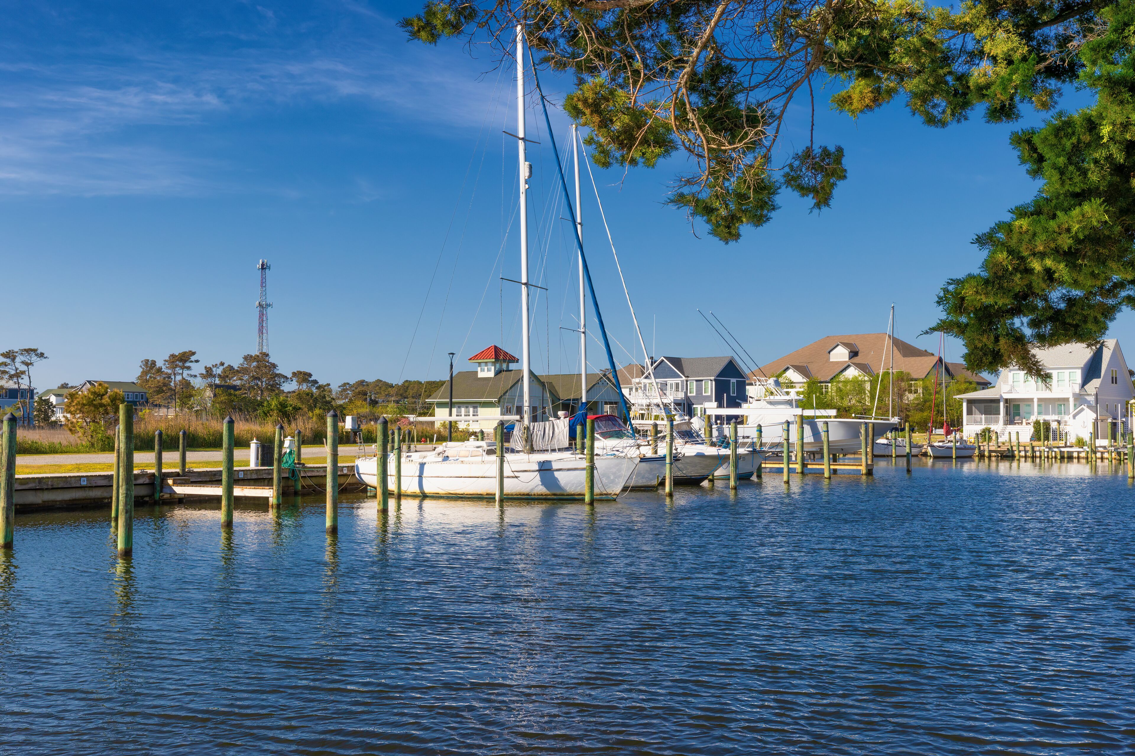 A marina with boats docked, a lighthouse in the background, and houses along the shore.