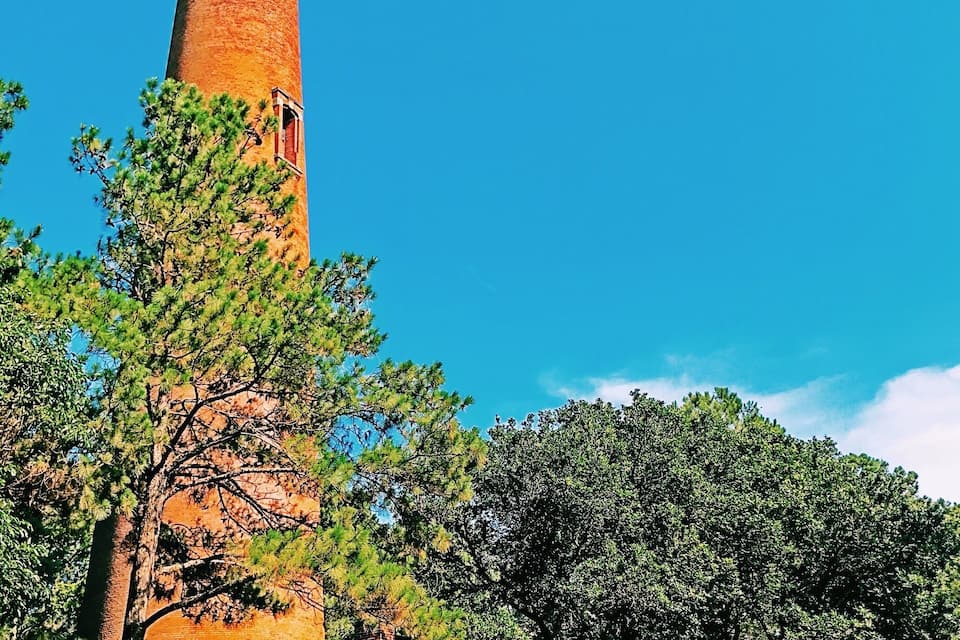 A tall brick lighthouse with a small house at its base, surrounded by trees and grass.