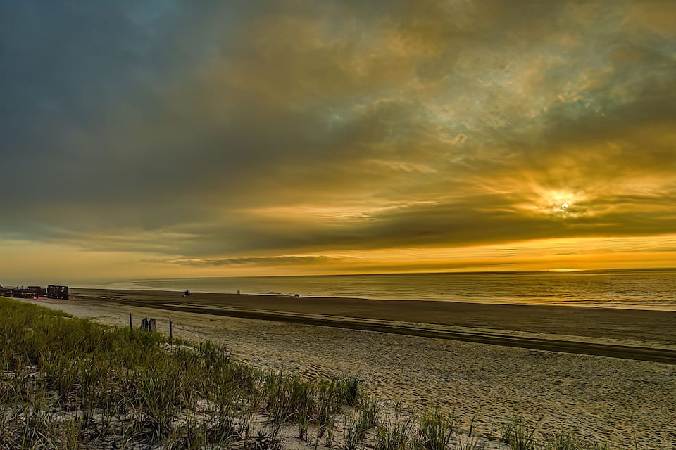 A beach with a sunset, grassy dunes, and a distant road.