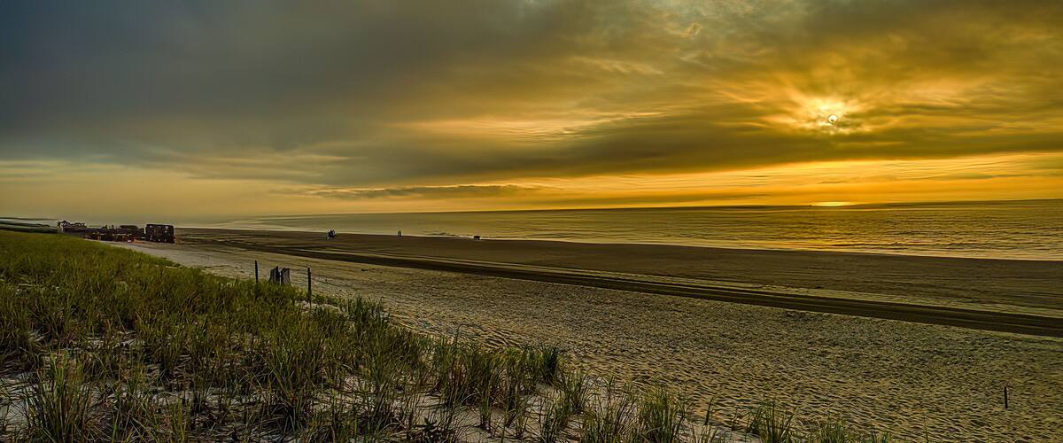 A beach with a sunset, grassy dunes, and a distant road.