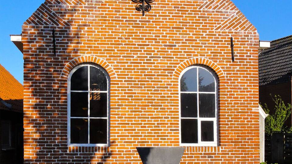 A small brick building with a tiled roof, two arched windows, and a decorative emblem above the entrance.