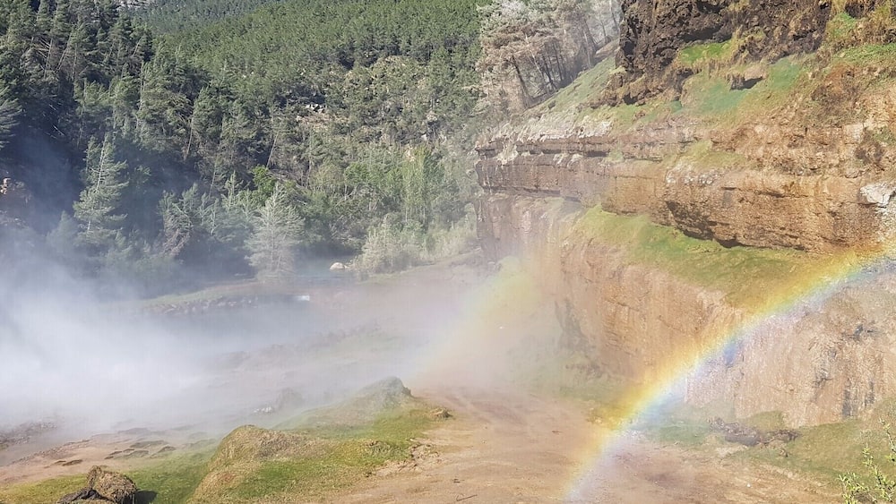A mountainous landscape with a visible rainbow, dense forest, and rocky cliffs.