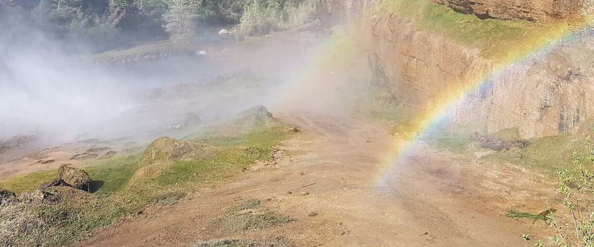A mountainous landscape with a visible rainbow, dense forest, and rocky cliffs.