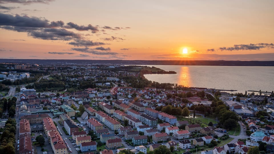 A coastal cityscape at sunset with residential buildings, a body of water, and a distant bridge.