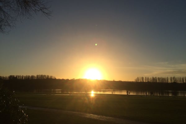 Un coucher de soleil sur un lac, avec les silhouettes d’arbres et de personnes qui se promènent.