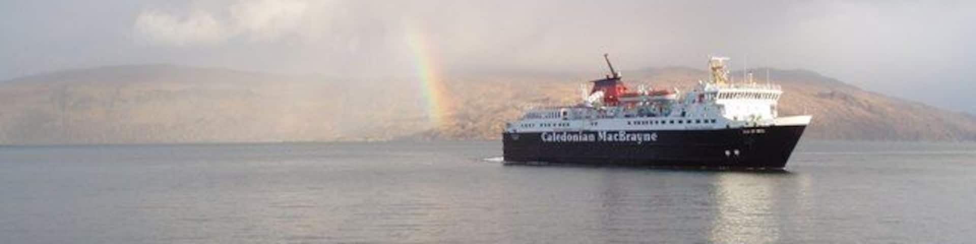 A ship named Caledonian MacBride on calm waters with a rainbow in the background and rocky shoreline in the foreground.