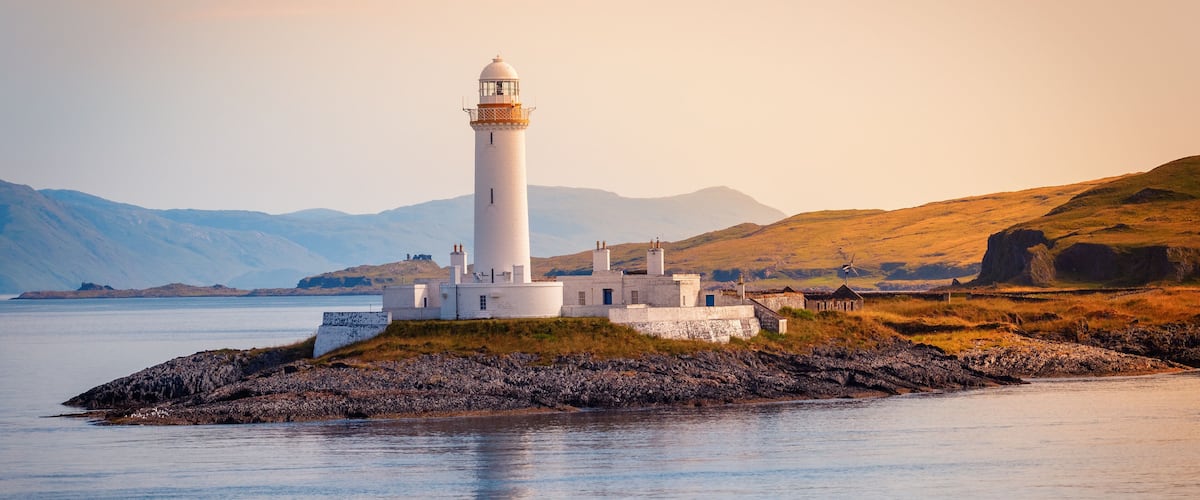 A lighthouse on a rocky island with calm water and distant mountains.