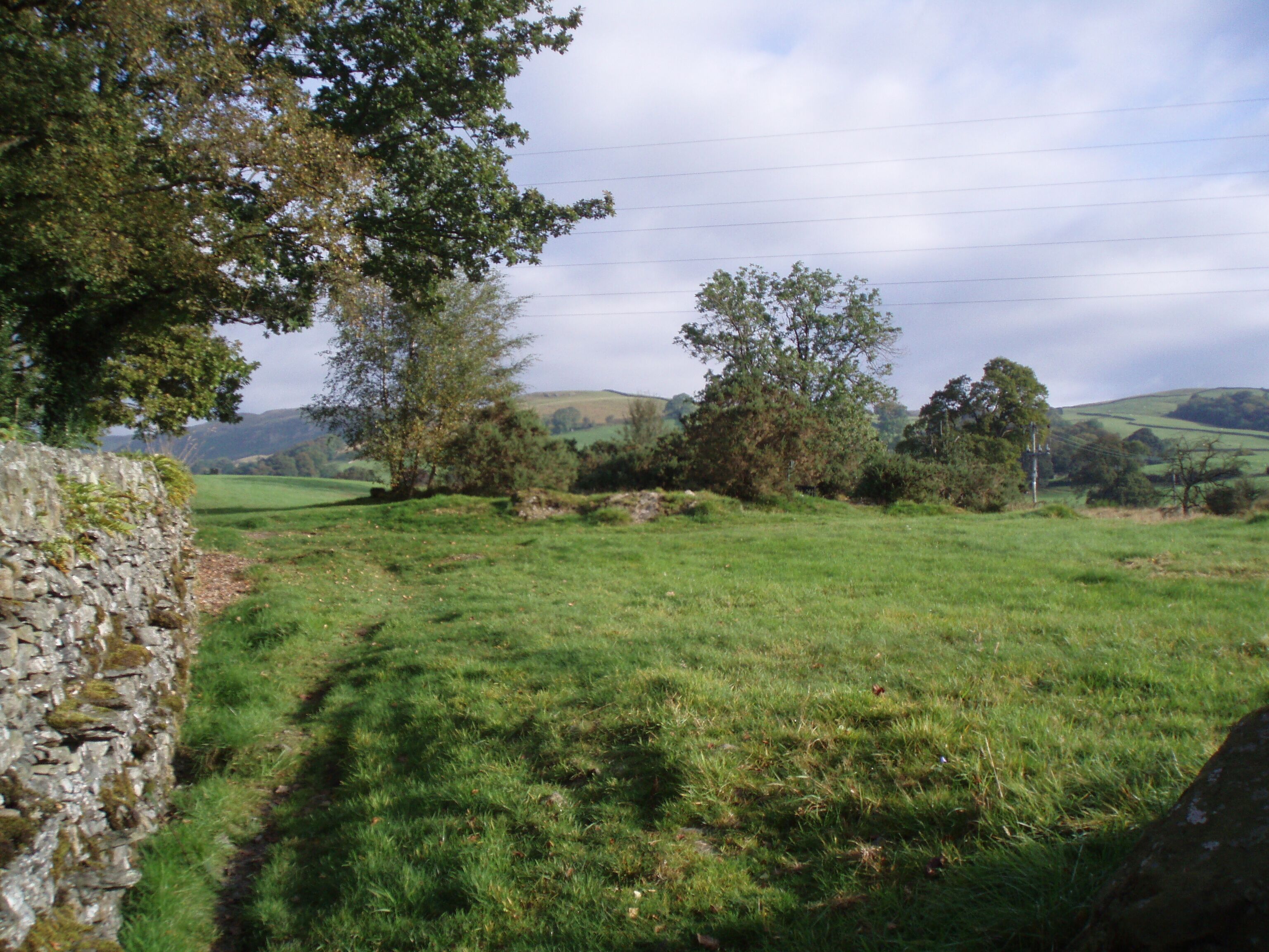A rural landscape with a stone wall, grassy field, trees, and rolling hills in the distance.