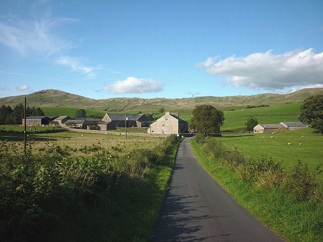 A rural road leading to a cluster of farm buildings in a hilly landscape.