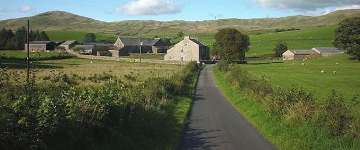 A rural road leading to a cluster of farm buildings in a hilly landscape.