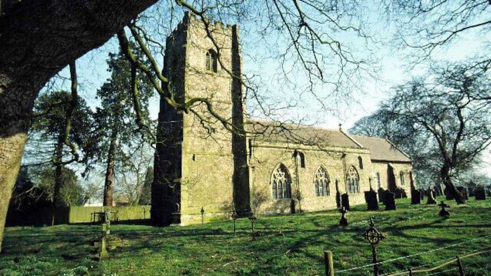 A church with a tall tower and arched windows, surrounded by a grassy field and trees.