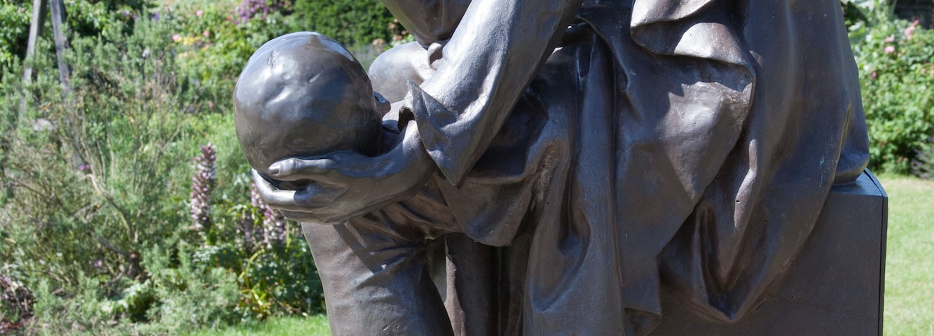 Estátua em bronze de um homem em pose pensativa, com vegetação e um céu claro ao fundo.