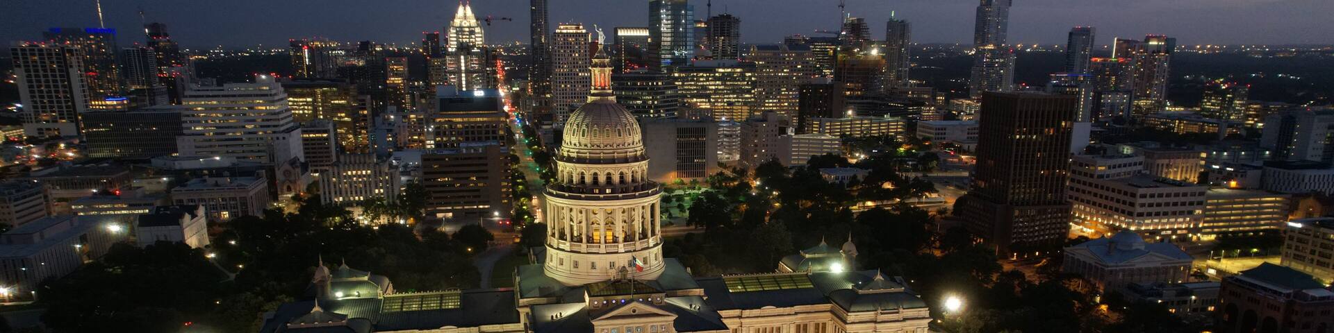 A cityscape at dusk with a prominent government building featuring a large dome, surrounded by modern skyscrapers and illuminated streets.