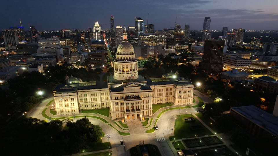 A cityscape at dusk with a prominent government building featuring a large dome, surrounded by modern skyscrapers and illuminated streets.