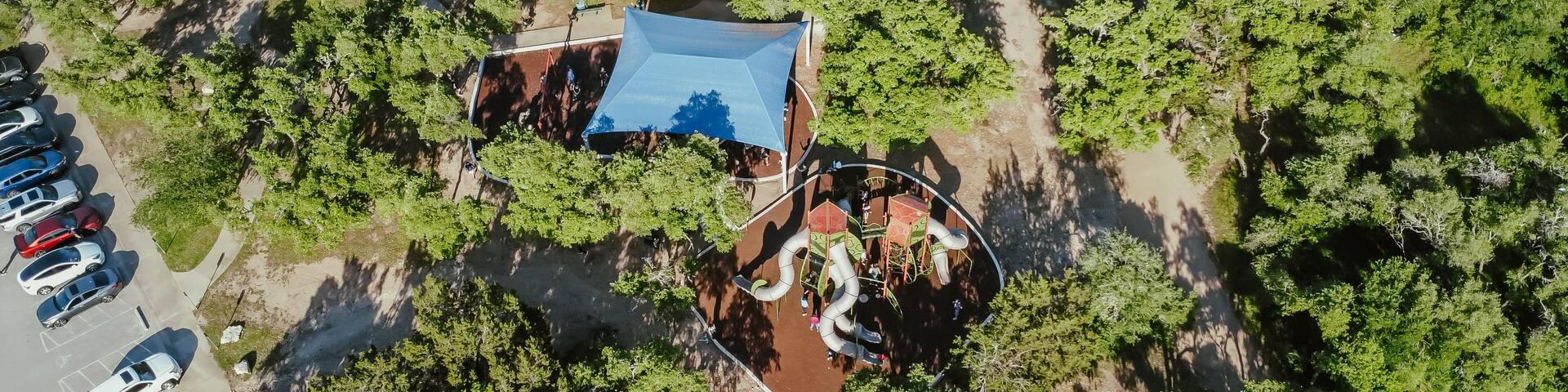 An aerial view of a park with a playground, a parking lot, and surrounding greenery.