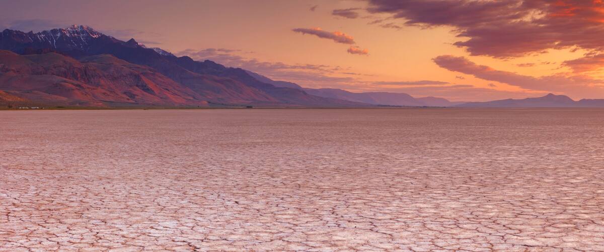 A vast, cracked desert landscape with mountains in the background and a sunset sky.