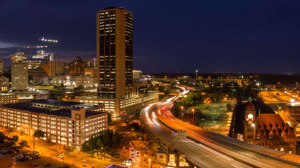 A nighttime cityscape with illuminated buildings, a prominent clock tower, and a curved highway.