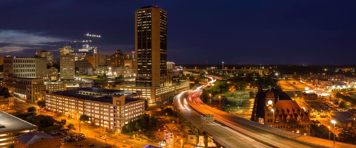 A nighttime cityscape with illuminated buildings, a prominent clock tower, and a curved highway.